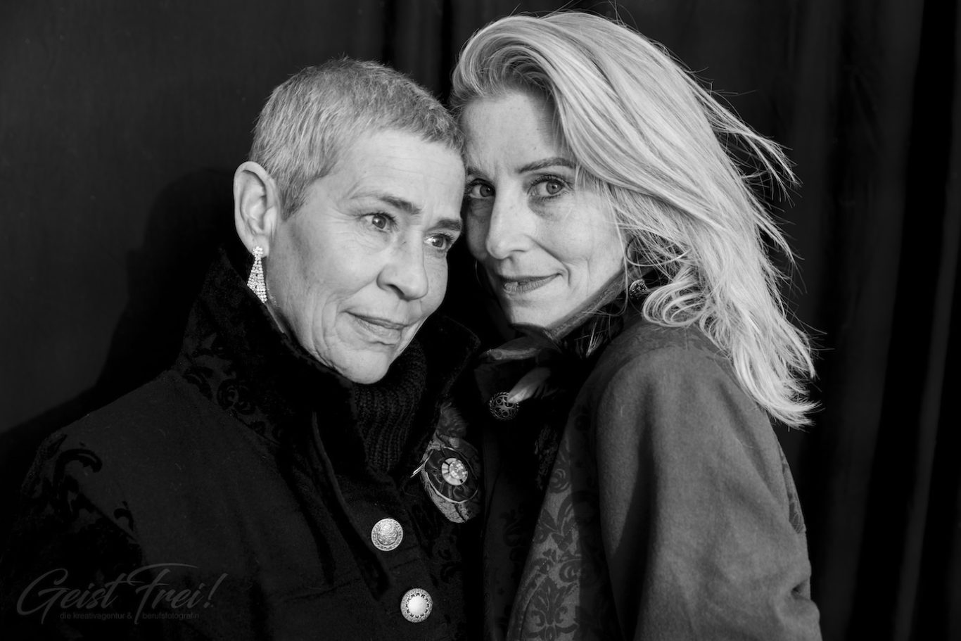 two Ladys Zwei Frauen posieren eng umarmt vor einem dunklen Hintergrund, eine mit kurzen grauen Haaren.