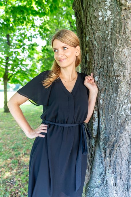 Junge Frau in schwarzem Kleid lehnt an einem Baum, umgeben von grünen Blättern.