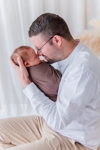 Vater hält schlafendes Baby in den Armen, beide lächeln sich an.