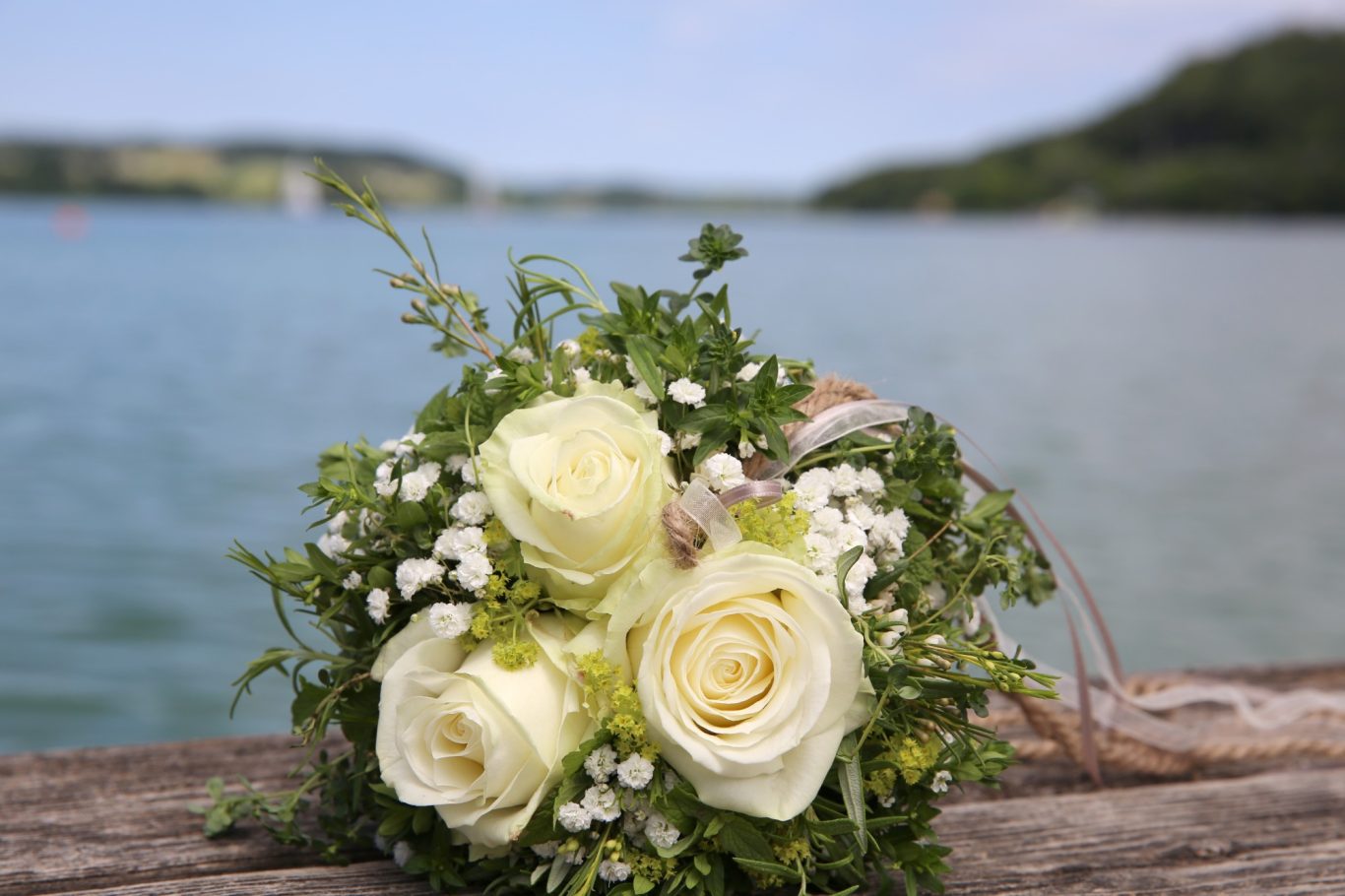 Brautstrauss Hochzeit GeistFrei! Ein Brautstrauß mit weißen Rosen und grünen Pflanzen vor einem Gewässer.