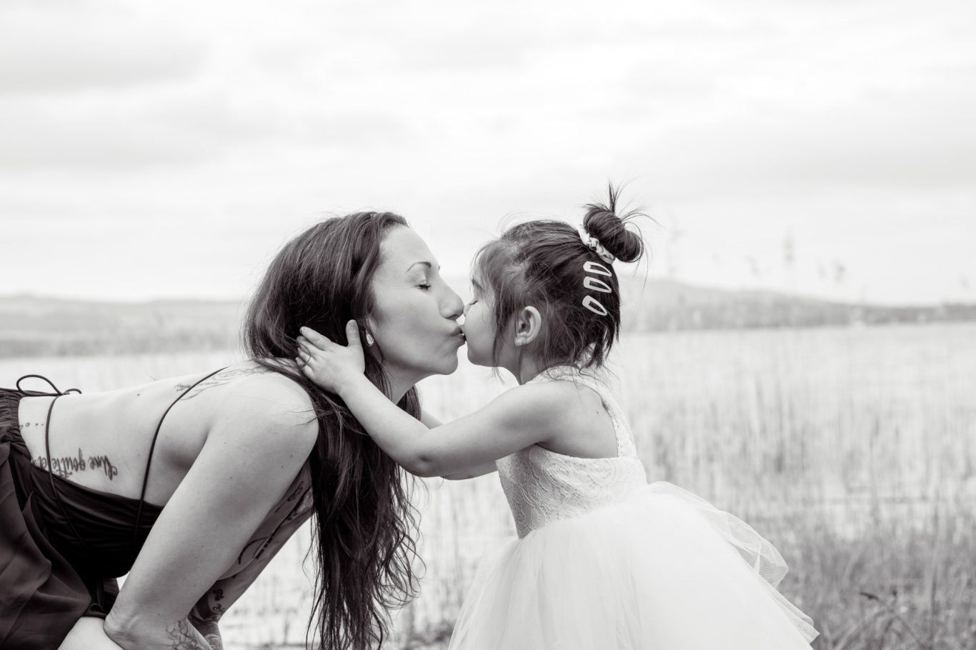 Mutter und Tochter küssend GeistFrei! Eine Mutter küsst ihre Tochter, beide lächeln glücklich vor einer natürlichen Landschaft.