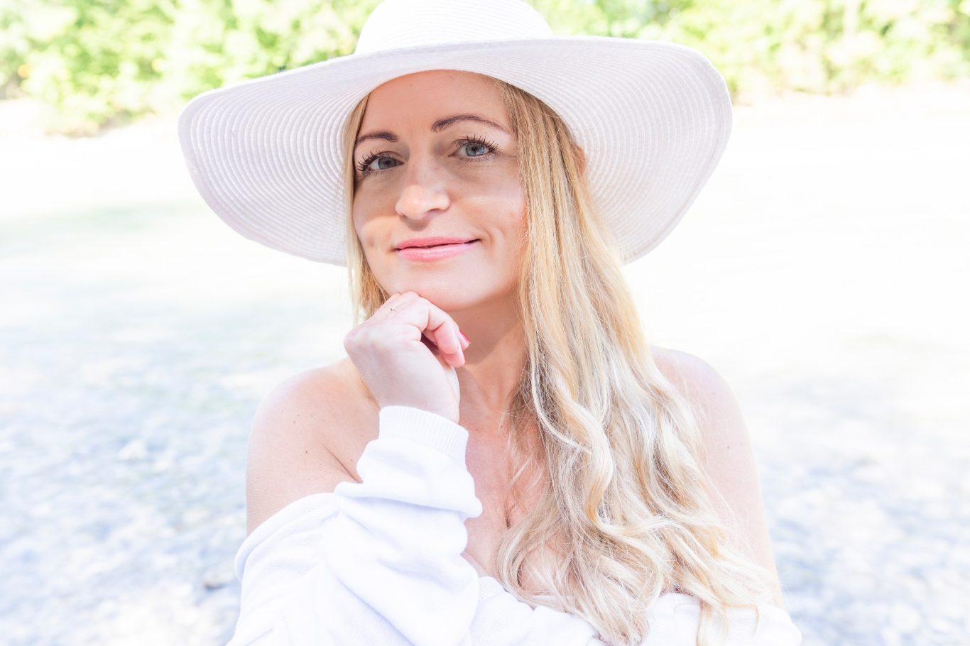 Blonde Frau mit Hut lächelt Blonde Frau mit Hut lächelt, mit einer Hand am Kinn, vor natürlichem Hintergrund.