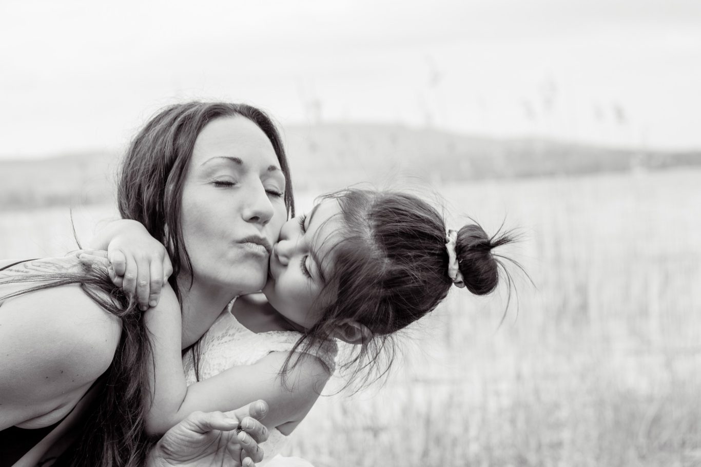 Mutter Tochter Liebe GeistFrei! Eine Mutter küsst ihre Tochter, beide lächeln glücklich in einer naturbelassenen Umgebung.