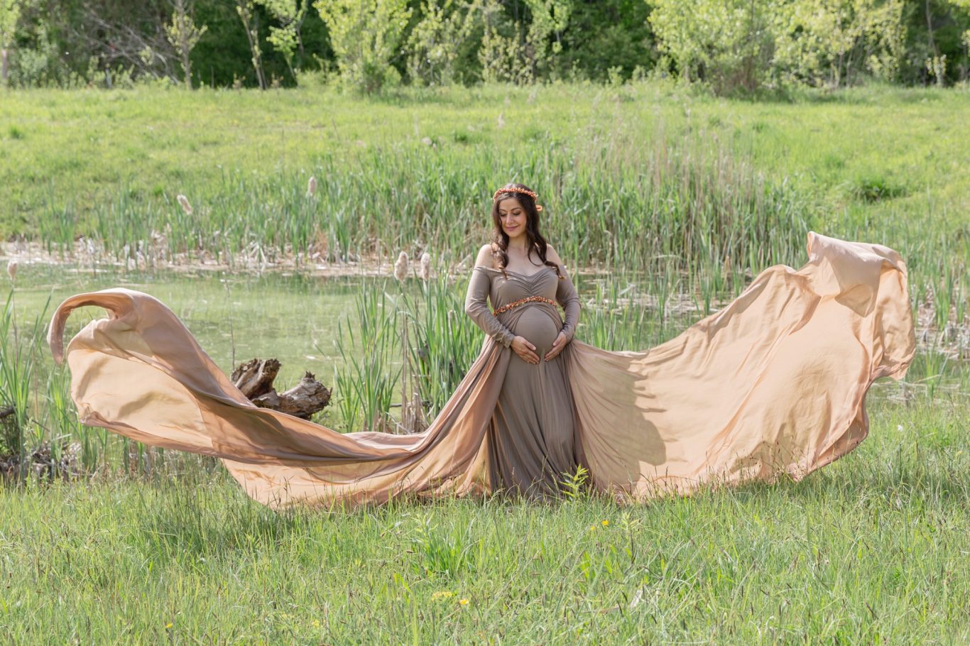 babybauchshooting babybauchfotografie werdende Mama GeistFrei Frau in einem fließenden, beigen Kleid steht in einer grünen Landschaft mit Wasser.