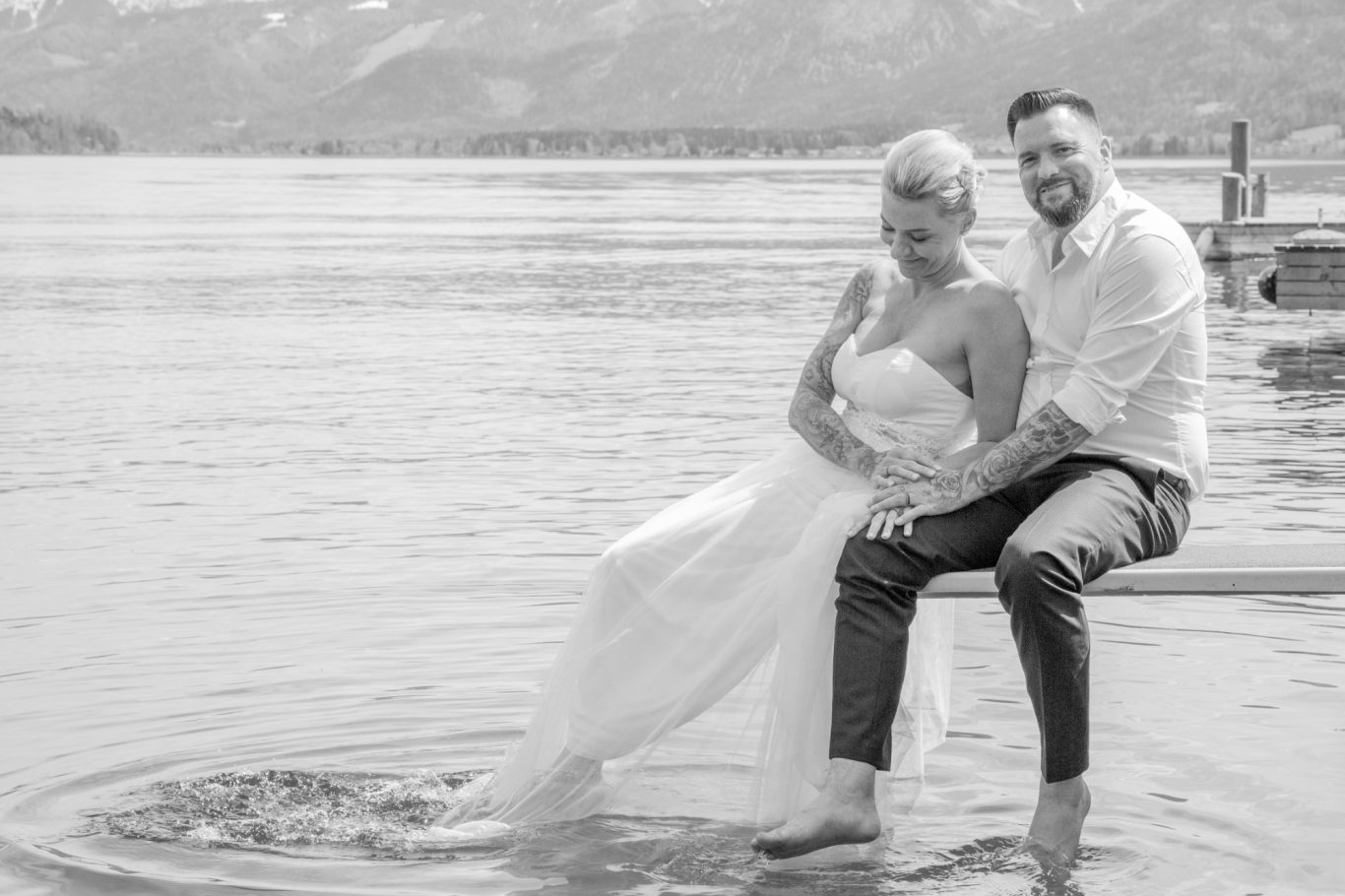 hochzeitsshooting, hochzeitsfotgrafie, wedding, trauung GeistFrei Ein Brautpaar sitzt am Ufer eines Sees, die Füße im Wasser und lächelt sich an.