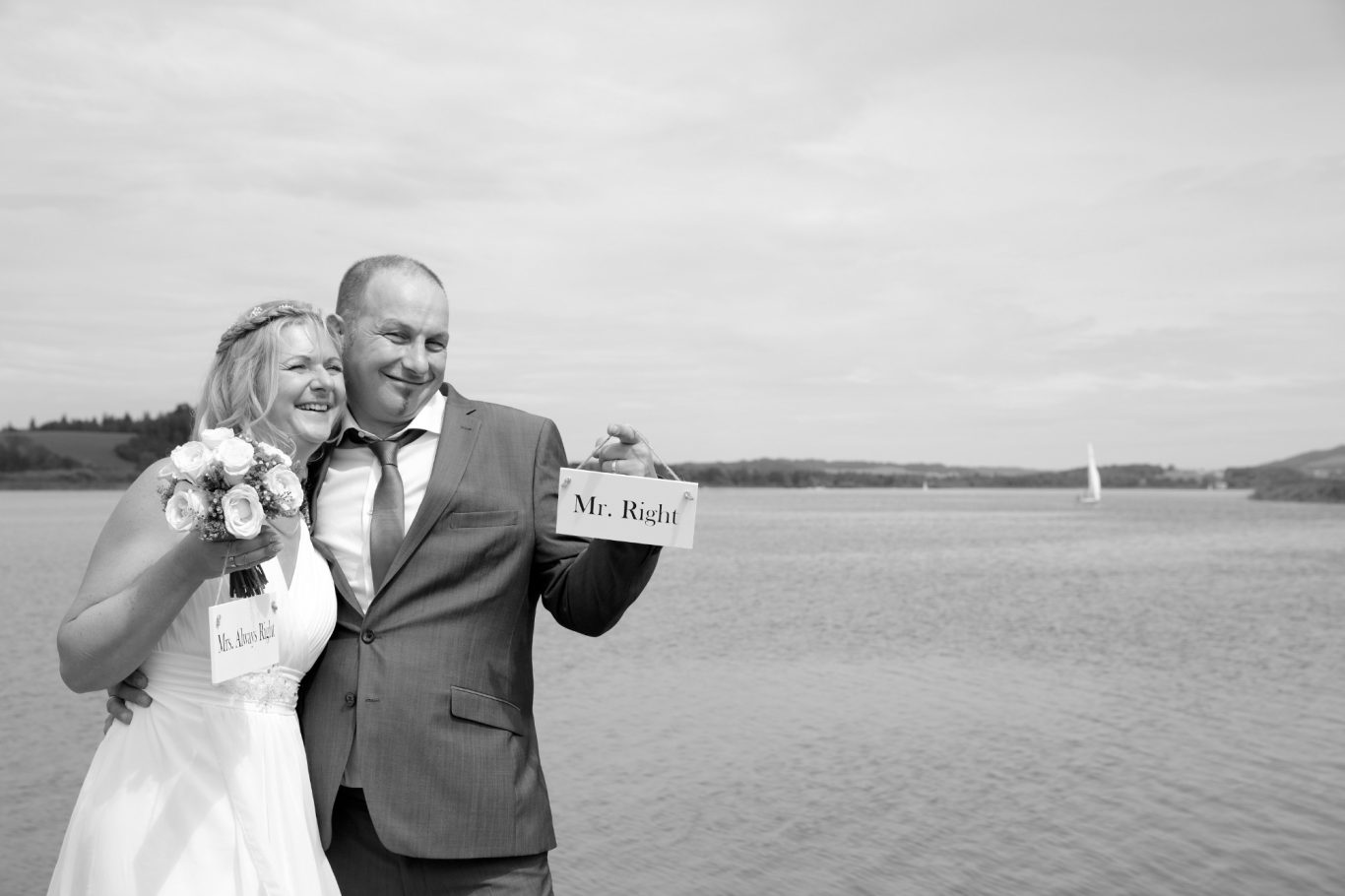hochzeitsshooting, hochzeitsfotgrafie, wedding, trauung GeistFrei Brautpaar lächelnd am Wasser, hält ein Schild mit der Aufschrift "Mr. & Mrs.".