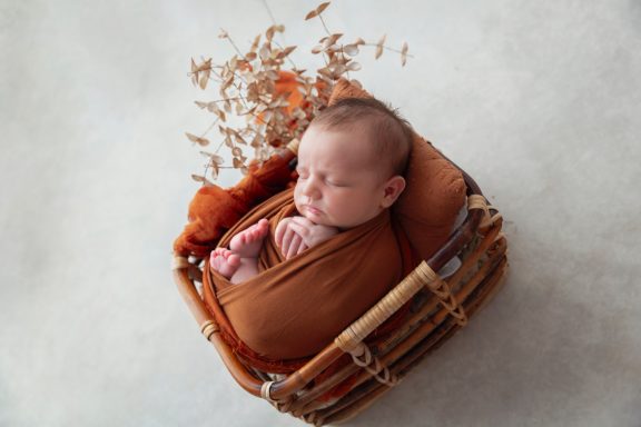 Neugeborenes Baby in einer geflochtenen Wiege, in braunes Tuch gehüllt, umgeben von Blättern.