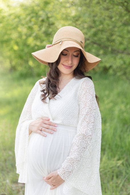 Schwangere Frau in weißem Kleid mit breiter Sonnenhat in einer grünen Wiese.