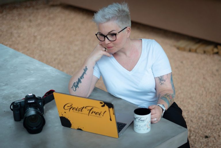 GeistFrei! die kreativagentur & berufsfotografin Ines Wolters-Pilz Frau mit kurzen Haaren sitzt an einem Tisch, Laptop und Kaffeetasse vor sich.