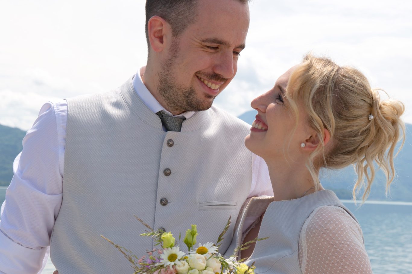 das glückliche Ehepaar Trauung GeistFrei! Brautpaar lächelt sich an, am Wasser mit einem Blumenstrauß in der Hand.
