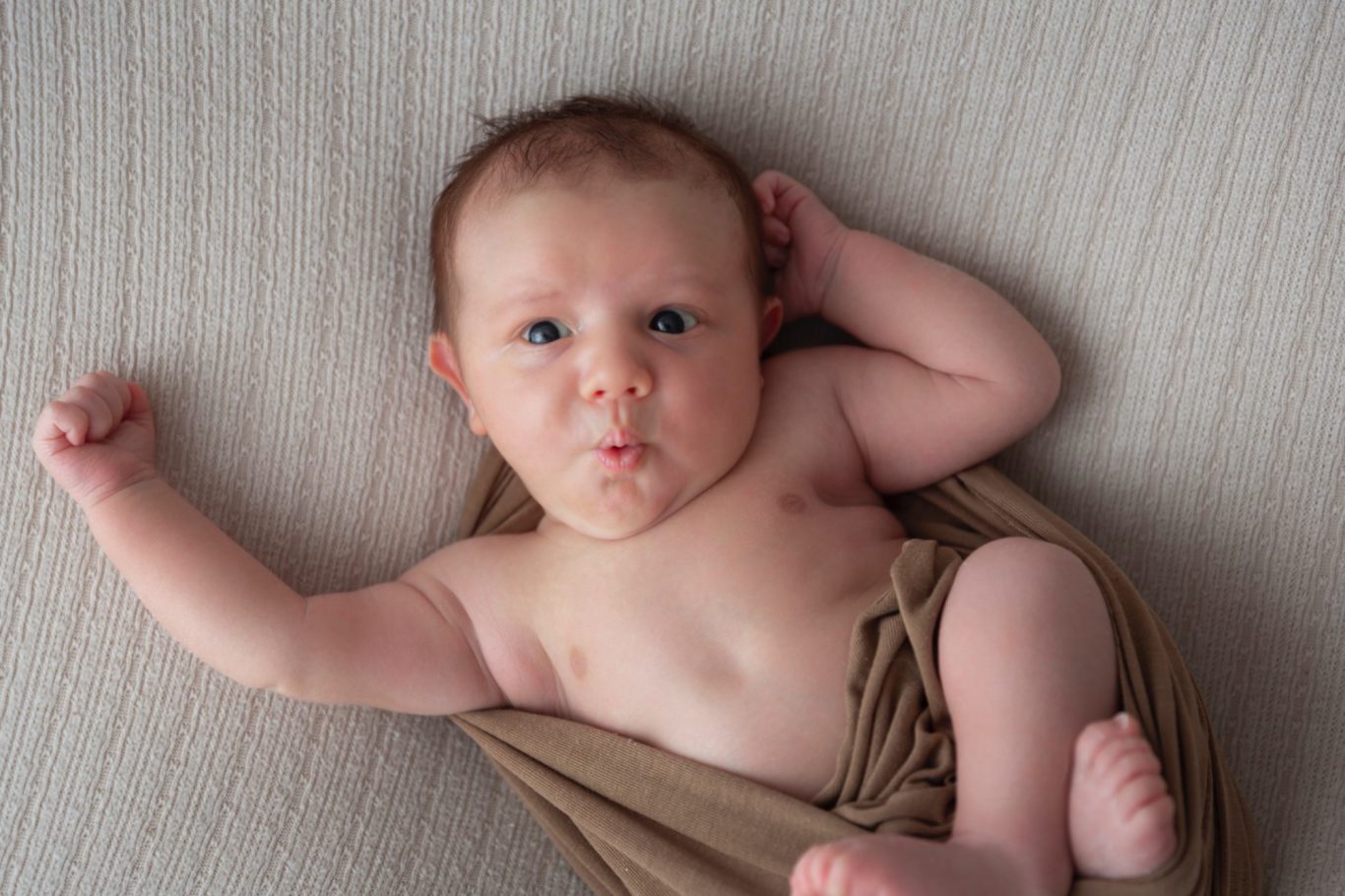 oh was ist denn los? newborn GeistFrei! Ein nacktes Baby liegt auf einer Decke, schaut mit leicht geöffnetem Mund neugierig.