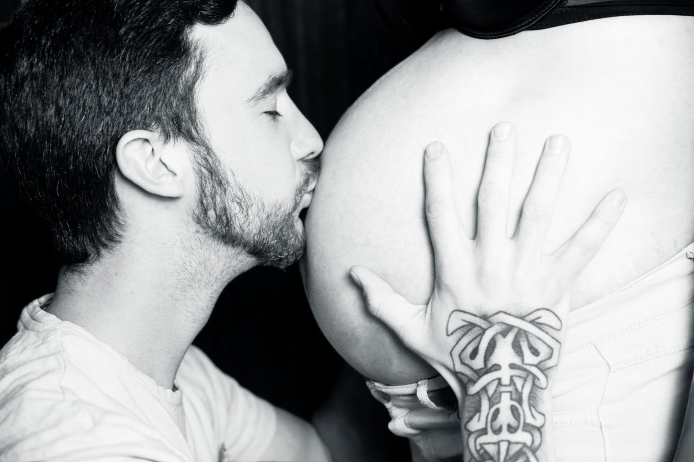 babybauchshooting babybauchfotografie werdende Mama GeistFrei Mann küsst einen runden Bauch, während er mit einer Hand darauf zeigt.