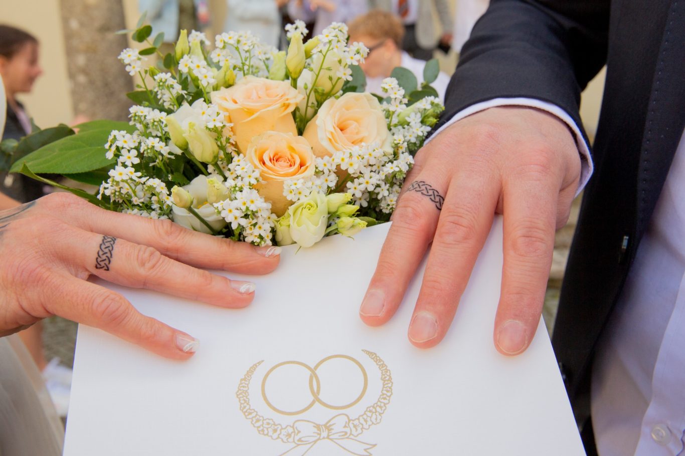 hochzeitsshooting, hochzeitsfotgrafie, wedding, trauung GeistFrei Hände mit Trauringen halten eine Hochzeitskarte und einen Blumenstrauß.