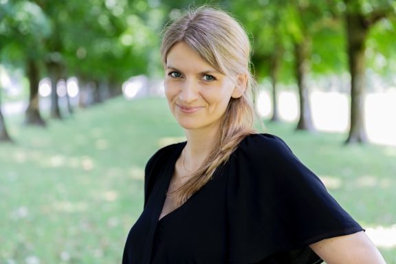 Frau mit blonden Haaren lächelt in einem Park mit Bäumen im Hintergrund.