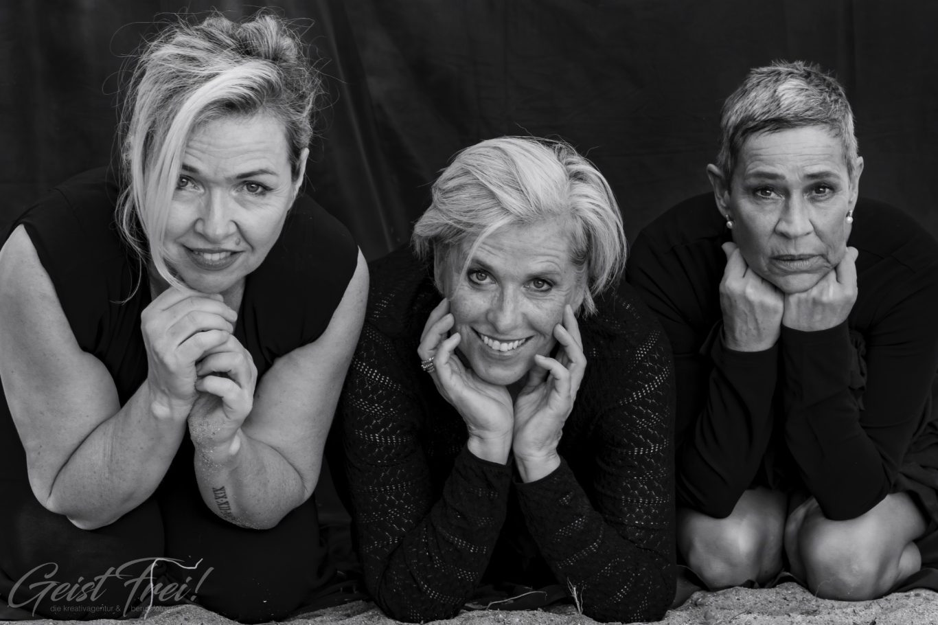 drei Ladys over 50 Drei Frauen in Schwarz-Weiß, lächelnd und mit Händen am Kinn, auf dem Boden sitzend.