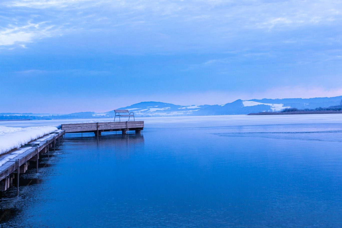 seekirchen am wallersee im winter Blick auf einen ruhigen See mit einem Holzsteg und verschneiten Bergen im Hintergrund.