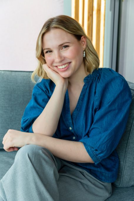 Lächelnde junge Frau mit blonder Haarfarbe in blauer Bluse auf einem Sofa.