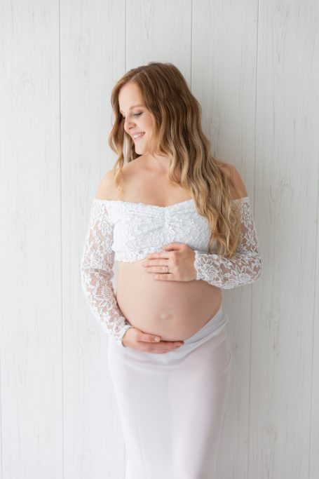 Schwangere Frau in weißem Kleid, lächelnd und den Bauch haltend vor einer hellen Wand.