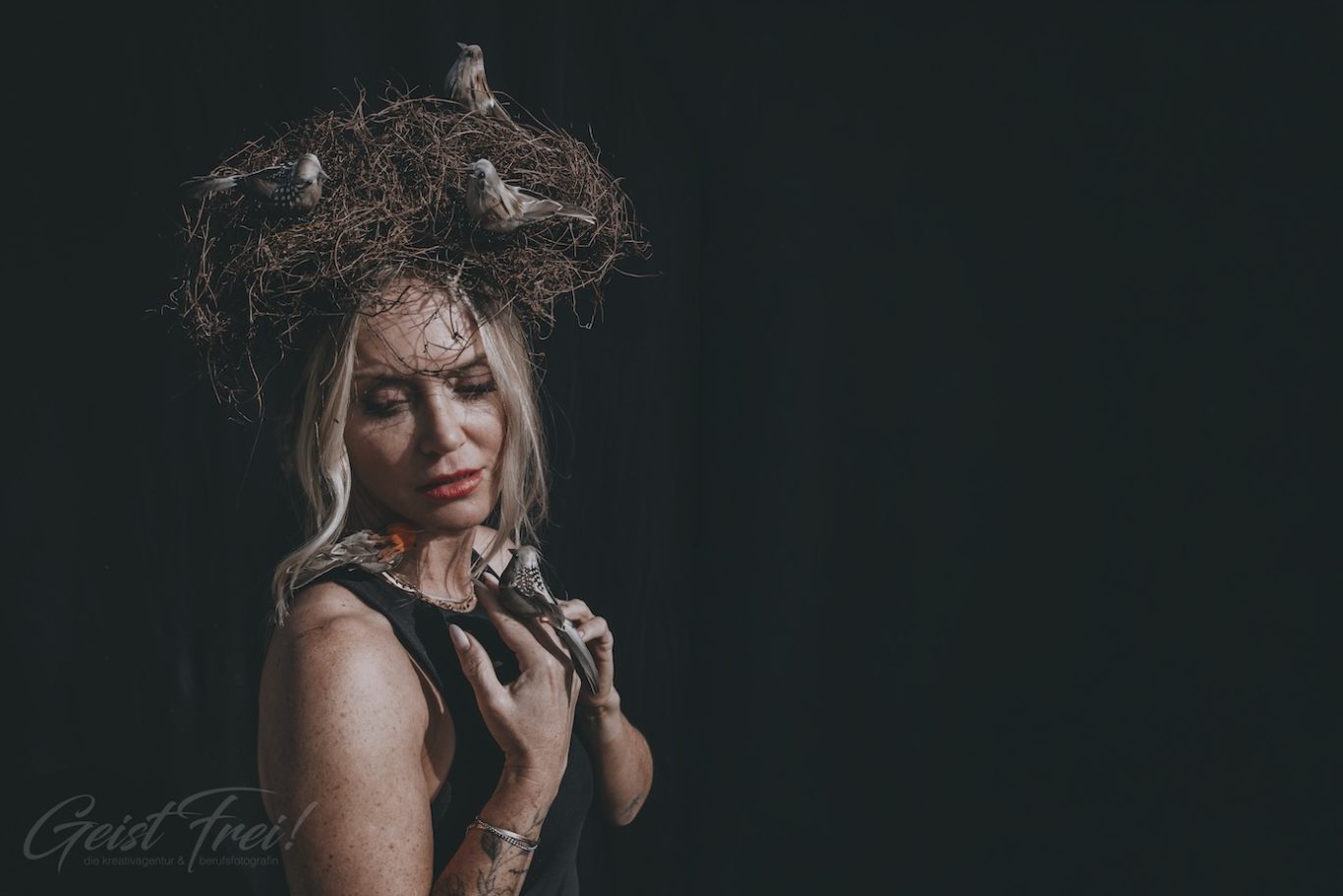 Vogelnest melancholisch Frau mit Vögeln am Kopf und elegantem Outfit vor dunklem Hintergrund.